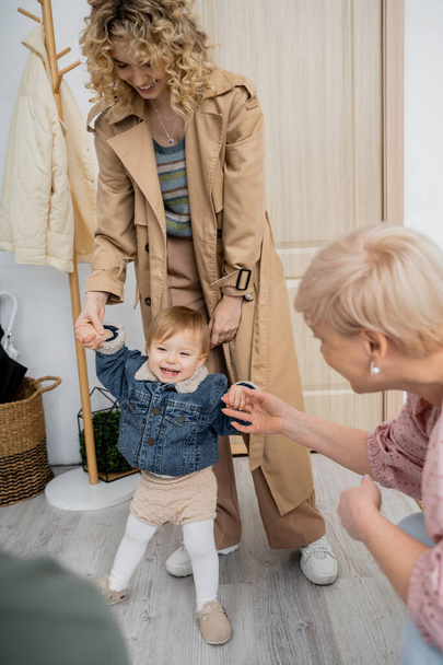 toddler girl in denim jacket smiling near mother and granny in hall at home - Photo, Image