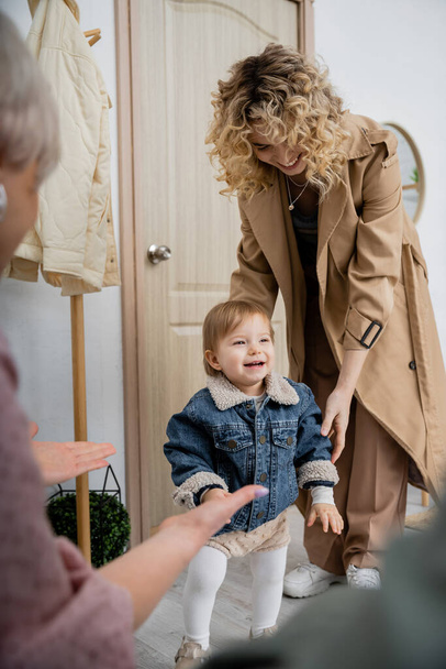 blurred woman meeting daughter with overjoyed child in denim jacket near door at home - Foto, afbeelding