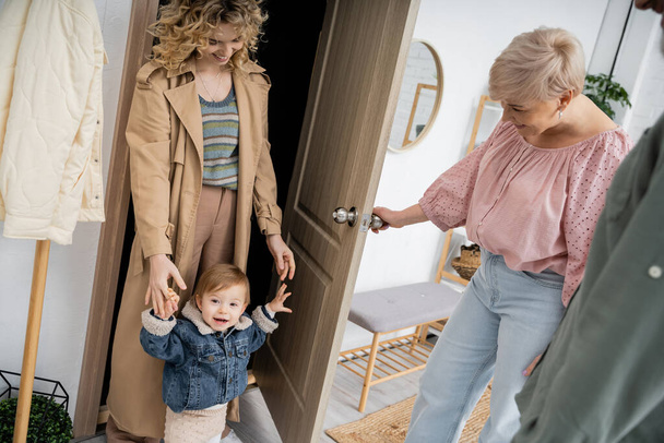 happy middle aged woman opening door to daughter with grandchild in denim jacket during their visit at home - Fotografie, Obrázek