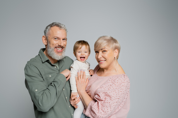glückliches älteres Paar blickt in die Kamera neben lachender Enkelin isoliert auf grau - Foto, Bild