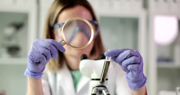 Lady scientist looks at dark transparent substance in test tube through magnifier glass. Concentrated woman examines state of liquid in glassware - Footage, Video