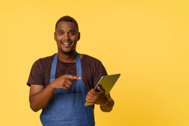 Young, talented African American chef search for the perfect recipe on his trusty tablet. Clad in a stylish blue apron isolated on yellow background, he symbolizes the fusion of modern technology and - Фото, изображение