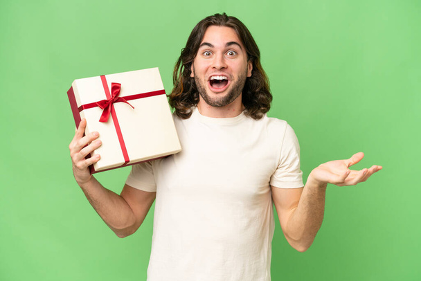 Jeune homme beau tenant un cadeau sur fond isolé avec expression faciale choquée - Photo, image