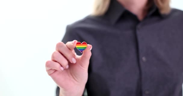 Mujer sostiene la insignia del arco iris en forma de corazón en la mano de pie sobre fondo blanco. Persona femenina con camisa gris muestra apoyo a la comunidad LGBT - Imágenes, Vídeo
