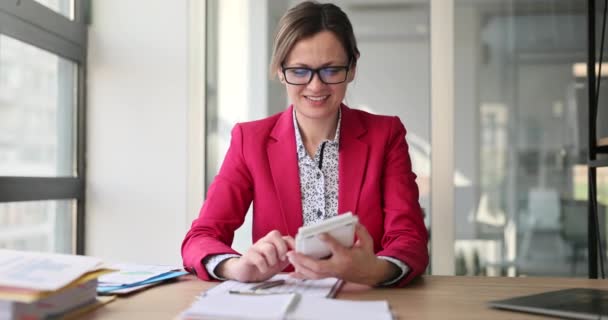 幸せな女性は会社の毎月の利益を計算した後の計算機で大きな数を示しています。レッドジャケットの感情的な会計士は仕事で喜びを感じますスローモーション - 映像、動画