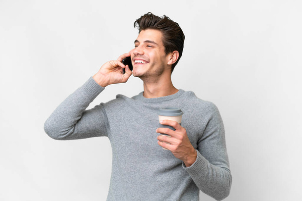 Young handsome caucasian man over isolated white background holding coffee to take away and a mobile - Photo, image