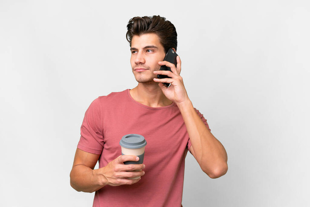 Young handsome caucasian man over isolated white background holding coffee to take away and a mobile - Фото, изображение