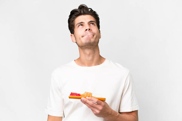 Young caucasian man holding sashimi over isolated white background and looking up - Fotó, kép