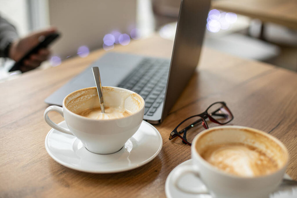 Portrait of coffee break with coffeespoon plate and having rest businessman in grey sweater typing someone on background. Black glasses laptop is on wooden table inside modern office.  - Foto, Bild