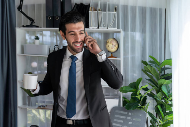 Diligent and busy businessman talking on phone call with coworker in modern office. Making sales call to client with coffee in hand, standing posing confidently as professional office worker. Fervent - Fotografie, Obrázek