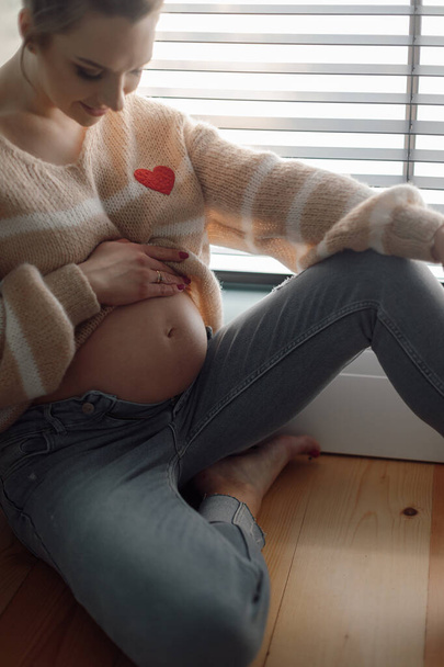 Чарівна молода вагітна жінка м'яко інсультує і оглядає шлунок, сидячи на підлозі біля вікна. Мати-дитина. Довгоочікувана вагітність. Чекаю на дитину. Радісні емоції
.  - Фото, зображення