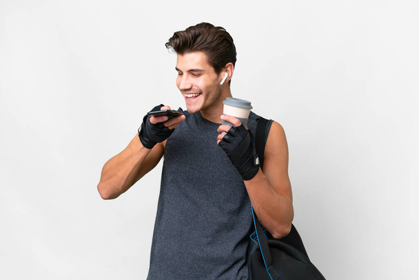 Joven hombre caucásico deporte con bolsa de deporte sobre fondo blanco aislado celebración de café para llevar y un móvil - Foto, Imagen