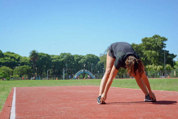 joven latina de etnia argentina estirándose en la pista de atletismo, de pie e inclinada hacia adelante tocándose los pies con las manos, entrenando preparándose para correr. - Foto, imagen