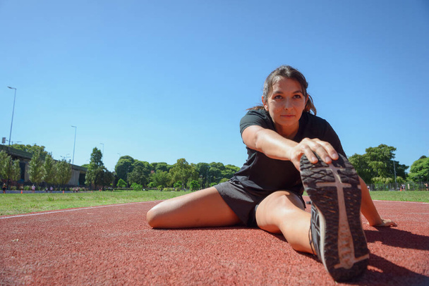 Wide angle front view of sporty young latina woman of Argentinian ethnicity, stretching and training on the running track, preparing to run and start her routine training, sport concept, copy space. - Photo, Image