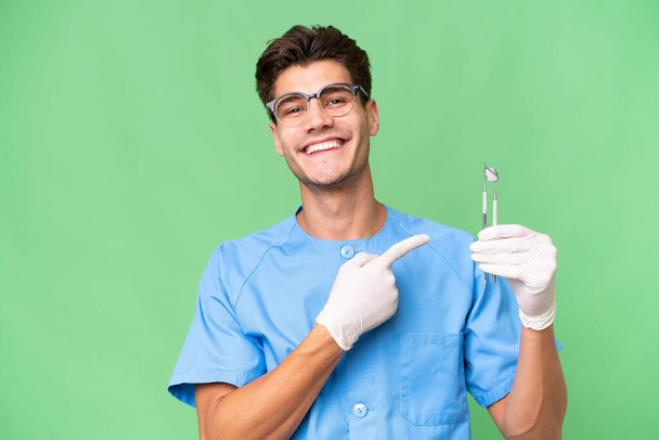 Jonge tandarts man met gereedschap over geïsoleerde achtergrond wijzend naar de zijkant om een product te presenteren - Foto, afbeelding