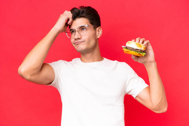 Mladý běloch drží burger izolovaný na červeném pozadí s pochybnostmi a zmateným výrazem ve tváři - Fotografie, Obrázek