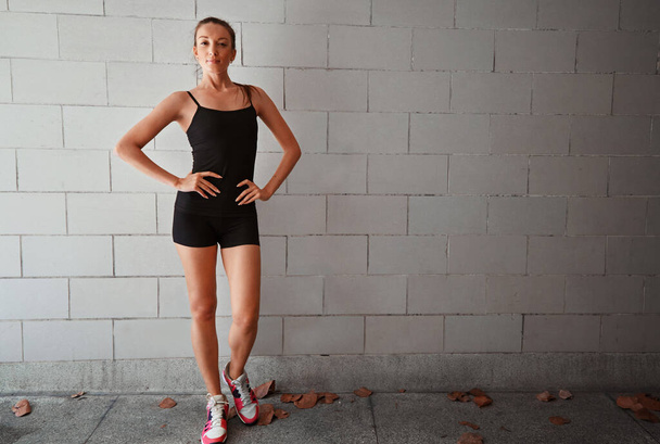 Давайте серьезно поговорим о здоровье. Портрет спортивной молодой женщины, стоящей с руками на бедрах у стены - Фото, изображение
