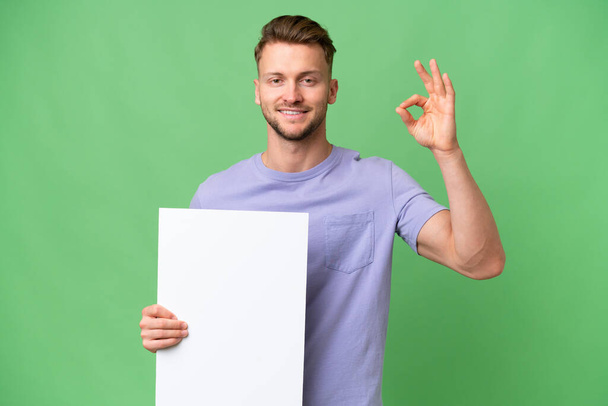 Young blonde caucasian man over isolated background holding an empty placard and doing OK sign - Foto, immagini