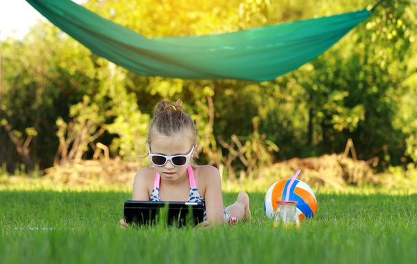 niña está comiendo pedazo de pizza y jugando en el ordenador portátil tumbado en la hierba en verano día soleado. el niño come después de los juegos activos. hamaca y bola en el fondo - Foto, imagen