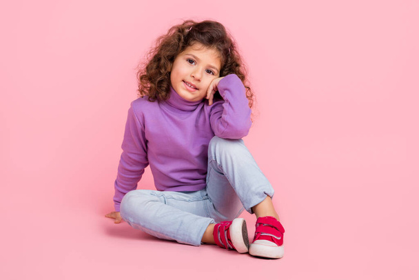 Повна довжина фото фанк-чарівна маленька дитина одягнена в фіолетовий пуловер, що сидить на підлозі, усміхаючись ізольований рожевий колір фону
. - Фото, зображення