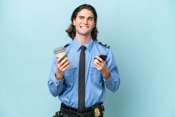 Joven policía caucásico aislado sobre fondo azul sosteniendo café para llevar y un móvil mientras piensa algo - Foto, imagen