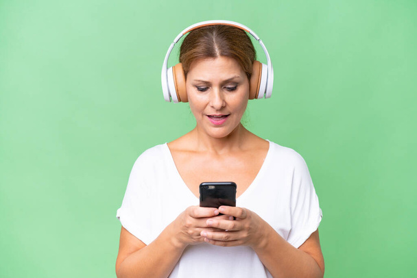 Donna caucasica di mezza età su musica d'ascolto di sottofondo isolata e guardando al cellulare - Foto, immagini