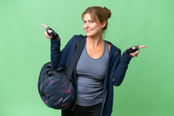Middle-aged sport woman with sport bag over isolated background pointing finger to the laterals and happy - Valokuva, kuva