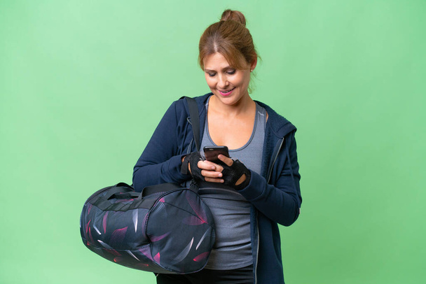 Sportlerin mittleren Alters mit Sporttasche über isoliertem Hintergrund sendet eine Nachricht mit dem Handy - Foto, Bild