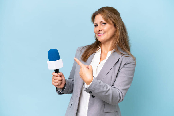 Middle-aged TV presenter woman over isolated blue background pointing back - Photo, Image
