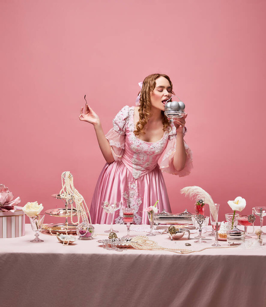Celebrating life. Fairy tale blond princess wearing renaissance dress standing near the laid festive table with ice cream over pink studio background. Concept of medieval, beauty, party, baroque, ad - Photo, Image