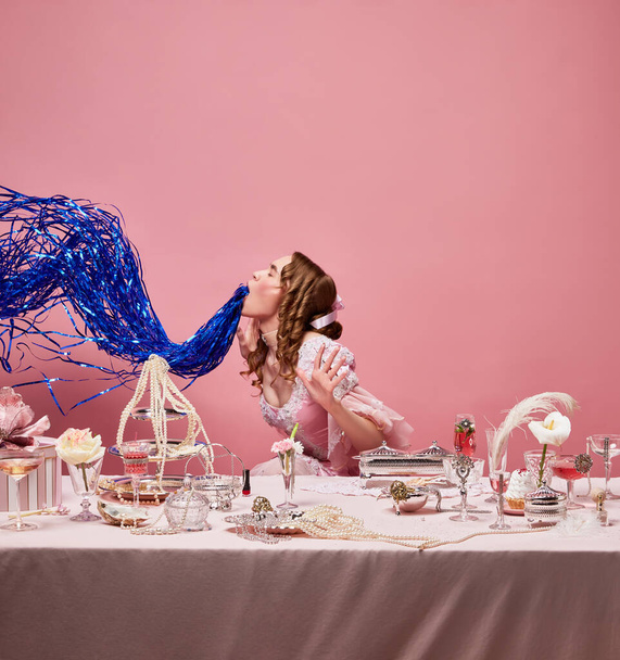 Alkoholová párty. Středověká atraktivní blond královna v nóbl růžové šaty sedí u stolu s kocovinou přes studio pozadí. Concept og oslava, krása, móda, víkend, reklama - Fotografie, Obrázek