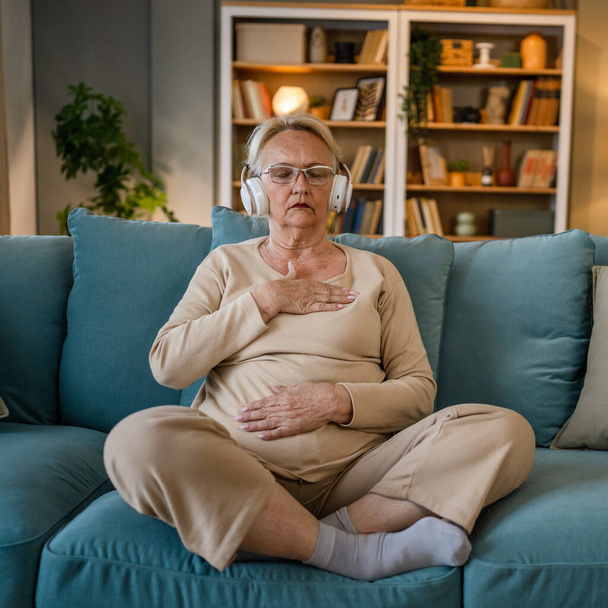 een vrouw volwassen senior Kaukasische vrouw met behulp van koptelefoon voor online geleide meditatie oefenen mindfulness yoga manifestatie met ogen dicht thuis echte mensen zelfzorg concept kopieer ruimte - Foto, afbeelding