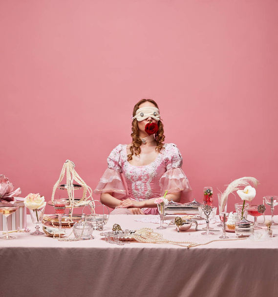 Banquet royal, rose rouge. Portrait de charmante reine portant un masque de sommeil, robe fantaisie et assise à la table allongée sur fond de studio rose. Concept de médiéval, nourriture, beauté, mode, annonce - Photo, image