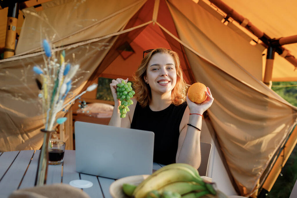 Lächelnde Freiberuflerin, die an einem sonnigen Tag am Laptop im gemütlichen Glamping-Zelt arbeitet. Luxus-Zeltlager für Sommerurlaub und Urlaub im Freien. Lifestyle-Konzept - Foto, Bild