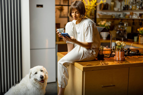 Young woman uses smartphone and drinks a coffee while sitting with her dog on kitchen at home. Concept of morning routine and domestic lifestyle - Foto, imagen