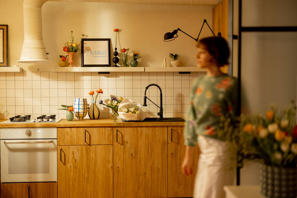 Moderno interior de cocina realizado en materiales naturales con fachadas de madera y estantes abiertos decorados con flores. figura femenina borrosa movimiento delante - Foto, Imagen