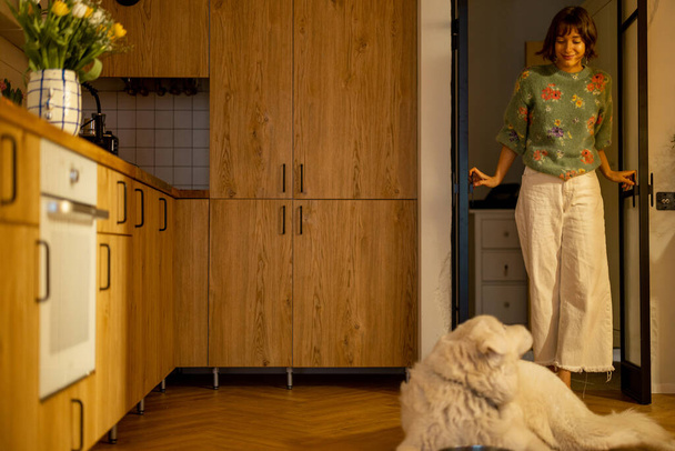 女性は台所の部屋を歩き、スタイリッシュな家のインテリアに横たわっている彼女の犬を迎えます。家庭生活の概念 - 写真・画像