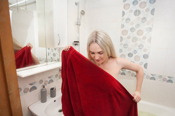A blonde woman wraps herself in a red bath towel, in the bathroom. Creative image on the theme of health, hygiene, taking care of your body. - Photo, Image