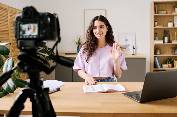 Attractive young woman sitting at desk in front of camera on tripod starting video for blog with greeting her followers - Photo, Image