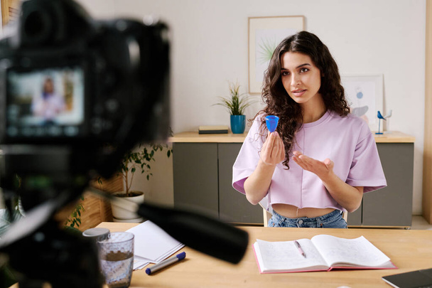 Junge Frau mit langen lockigen Haaren sitzt am Schreibtisch vor der Kamera und spricht über Erfahrungen mit Menstruationstasse, während sie ein Video für den Blog aufnimmt - Foto, Bild