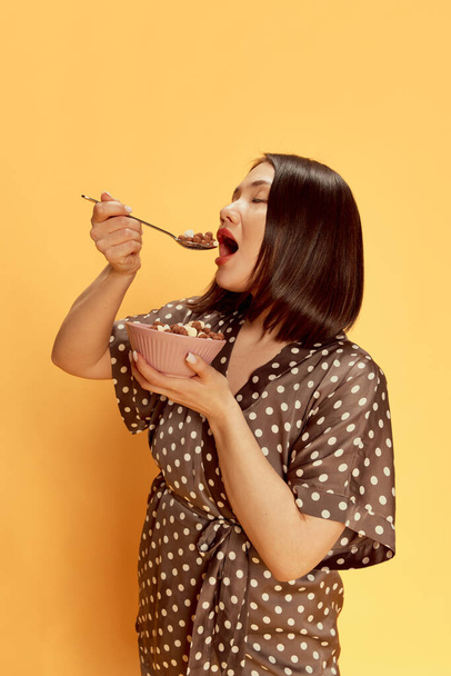 Portrait of beautiful, young, asian girl in stylish silk pajamas, eating cereal with milk against yellow studio background. Delicious breakfast. Concept of emotions, lifestyle, youth, comfort - Φωτογραφία, εικόνα