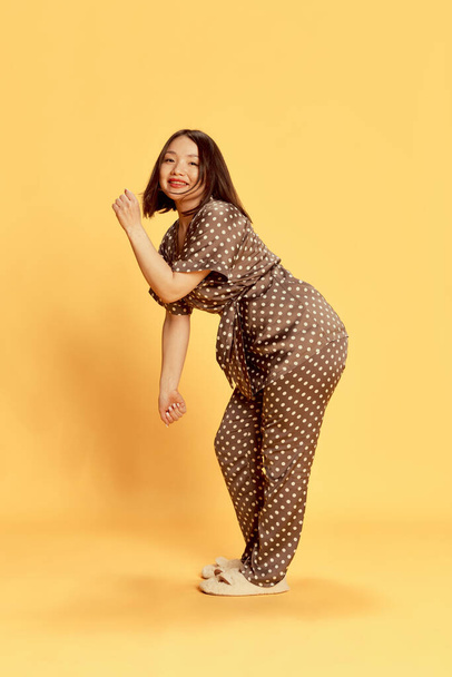 Full-length portrait of young, beautiful, asian girl in silk brown homewear, pajamas and slippers dancing against yellow studio background. Looking happy. Concept of emotions, lifestyle, youth - Fotoğraf, Görsel