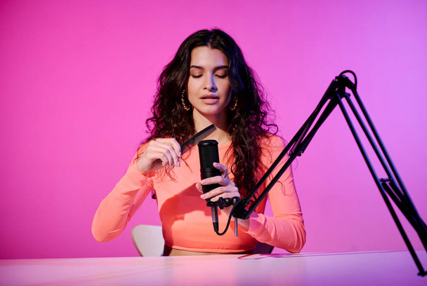 Elegante giovane influencer femminile caucasica seduta alla scrivania in studio registrazione di suoni realizzati con pettine per capelli per video ASMR, luce al neon rosa - Foto, immagini