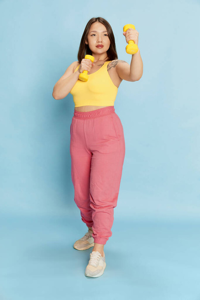 Full-length portrait of young asian girl in colorful, pink and yellow sportswear standing with dumbbells, training against blue studio background. Concept of emotions, healthy and sportive lifestyle - Photo, image