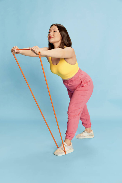 Full-length portrait of asian young girl, witch concentrated face, in comfortable, sportswear training with fitness expanders against blue studio background. Concept of emotions, lifestyle, youth - Φωτογραφία, εικόνα