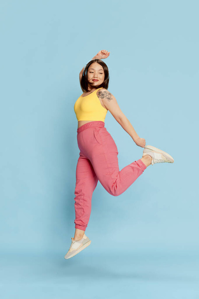 Full-length portrait of young, active, asian girl in colorful sportswear, listening to music in headphones, jumping against blue studio background. Concept of emotions, lifestyle, youth, sport - Foto, imagen