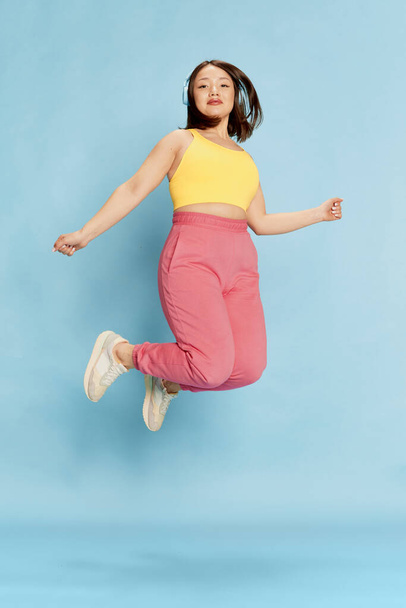 Full-length portrait of young asian girl in yellow top and pink trousers, listening to music in headphones and jumping against blue studio background. Concept of emotions, lifestyle, youth - Foto, Imagem