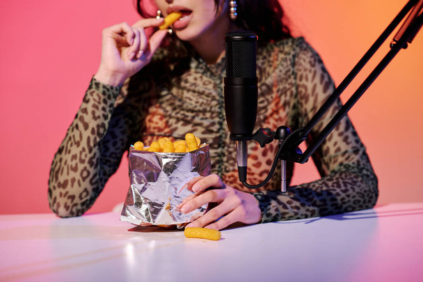 Stylish young female blogger sitting in studio in pink and yellow neon light recording corn puff snacks eating sounds for ASMR vlog - Foto, imagen