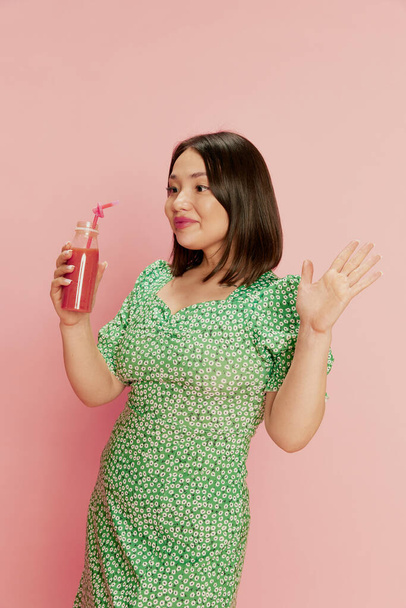 Portrait of positive, happy, young, asian girl in stylish green dress, drinking berry smoothie against pink studio background. Delicious food. Concept of emotions, lifestyle, youth, diet, health - Photo, image