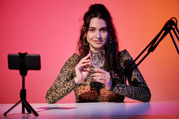 Élégante jeune femme caucasienne assise en studio en rose et orange néon enregistrement de sons et vidéo pour ASMR vlog en utilisant du verre de cristal - Photo, image